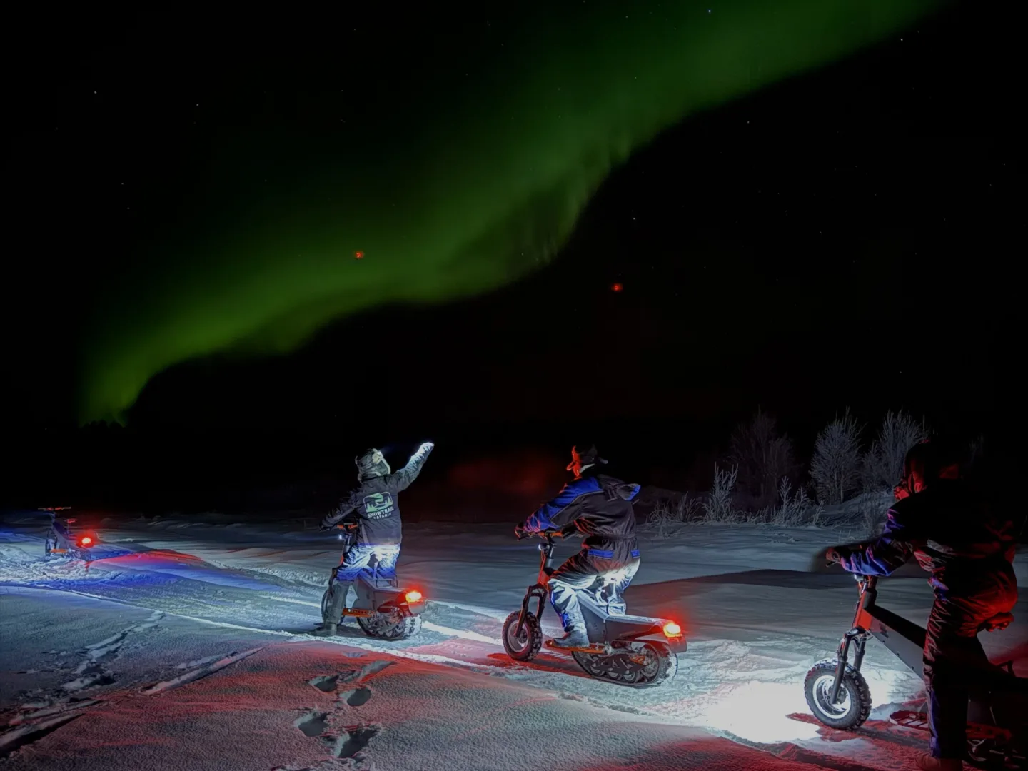Riders on electric snow scooters stopped under green northern lights at night