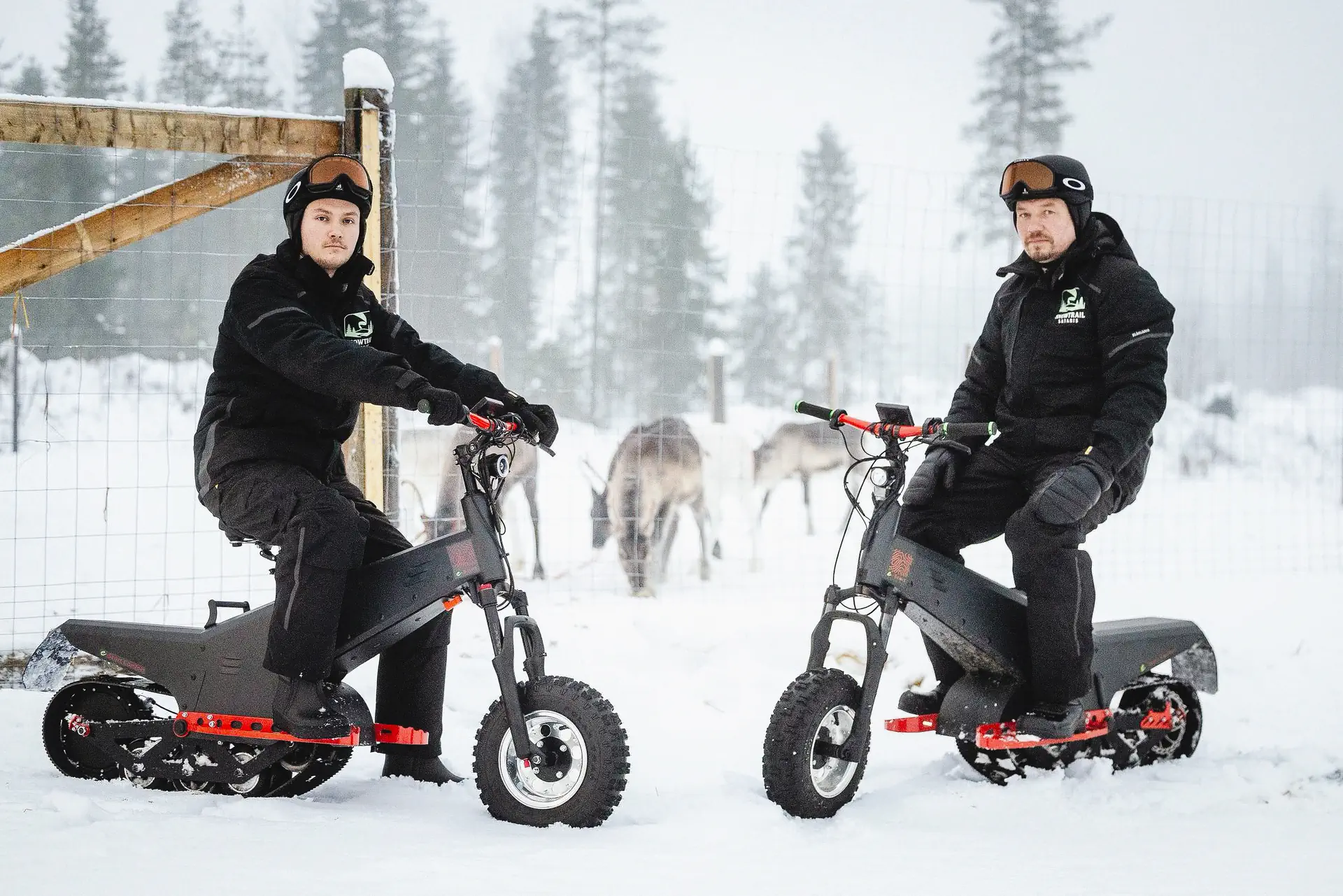 Due guide di Snowtrail Safaris su monopattini elettrici da neve accanto a un recinto di renne in un bosco innevato