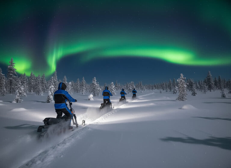 Where to See Northern Lights in Rovaniemi 2 Snowmobile group riding across open snow under bright green northern lights